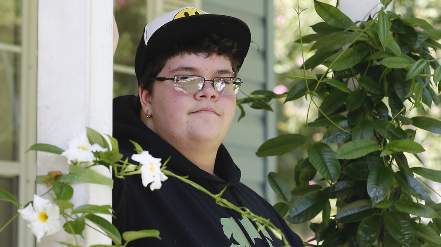 A teenage boy stands on a porch behind flowers.